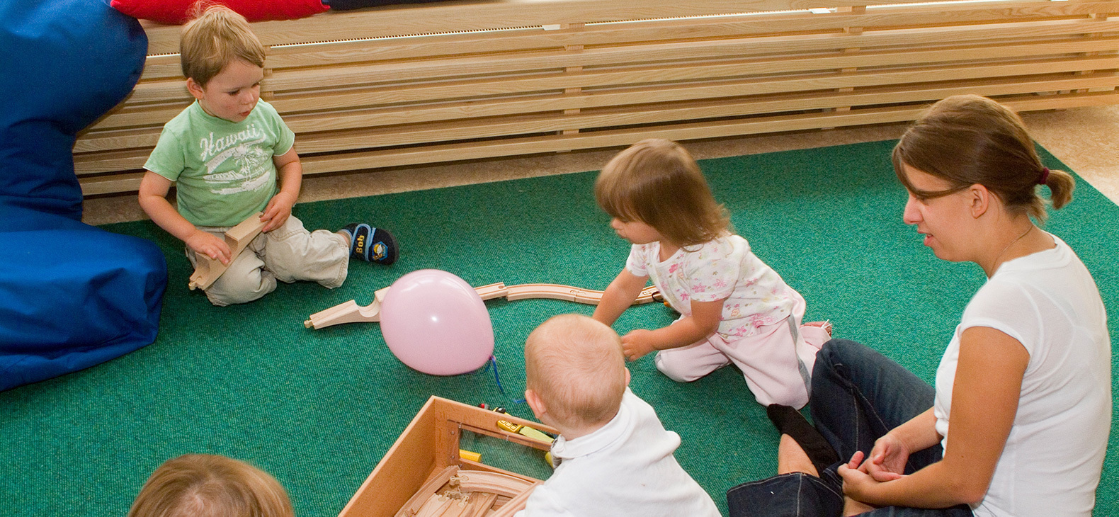 Eine Kindergärtnerin spielt mit vier kleinen Kindern auf dem Boden mit einer Holzeisenbahn und einem Luftballon.