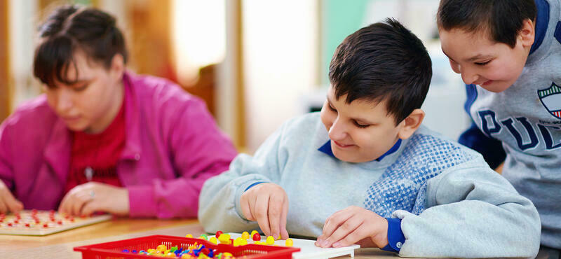 Ein Mädchen und ein Junge sitzen am Tisch und spielen mit Steckfiguren. Ein zweiter Junge schaut lächelnd zu.