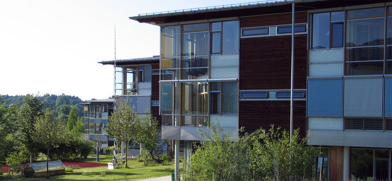 Außenfassade der kbo-Lech-Mangfall-Klinik Agatharied: Blick auf drei hintereinander angeordnete moderne Flachbauten mit Glas- und Holzelementen, auf einem parkähnlichen Gelände mit jungen Bäumen.