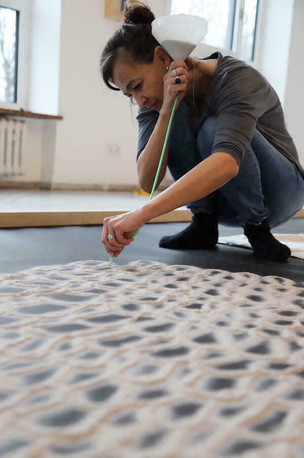 Frau kniet und erzeugt mit einem Schlauch an einem Trichter ein Muster am Boden aus feinen Sandspuren.