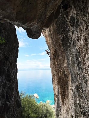Bergsteiger klettert an einer großen Höhlenbergwand nach oben. Im Hintergrund ist Meer zu sehen.