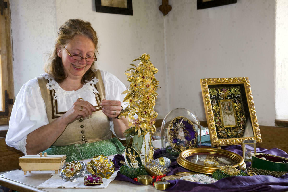 Eine Frau sitzt an einem Tisch, vor ihr viele filigrane religiöse Objekte, wie zum Beispiel ein Bilderrahmen.