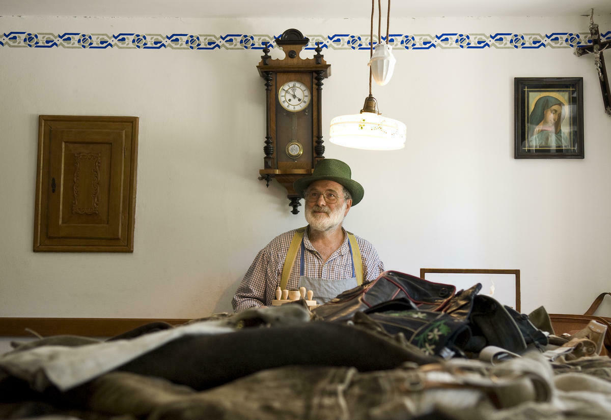 Ein Mann sitzt in einer alten Stube unter einer Uhr, vor ihm liegen auf einem Tisch Lederhosen.