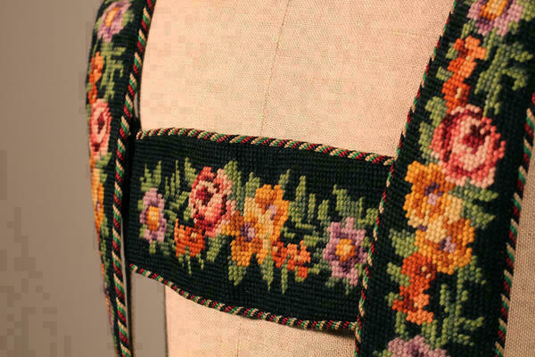 Detail eines mit Kreuzstichen bestickten Hosenträgers, das Muster bildet bunte Blumen und Blätter.