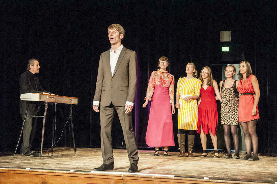 Szene auf einer Bühne: Ein Mann im Vordergrund singt, er wird begleitet von einem Keyboardspieler und einem fünfköpfigen weiblichen Backgroundchor.