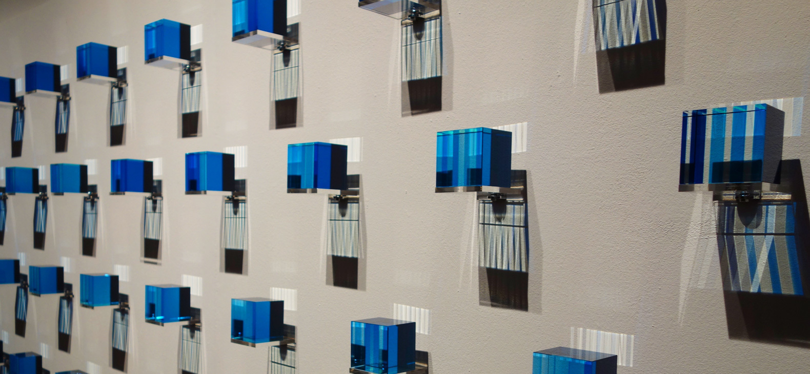 Blaue beleuchtete Plexigalswürfel werfen doppelte Schatten auf eine graue Wand.