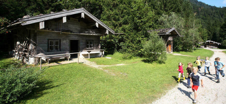 Das Holzknechtmuseum im Sommer: Auf einem Kiesweg laufen Kinder. Auf einer Wiese dahinter wirft das vorstehende Holz-Dach Schatten auf eine Block-Hütte. Dahinter steht dichter Nadelwald.