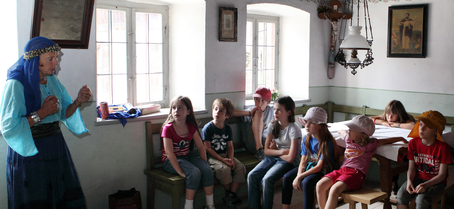 In einer geräumigen Stube in einem alten Bauernhaus: Eine Frau mit blauem Kopftuch und angehobenen Armen spricht zu einer Gruppe sitzender Kinder, die ihr gebannt Zuhörern.