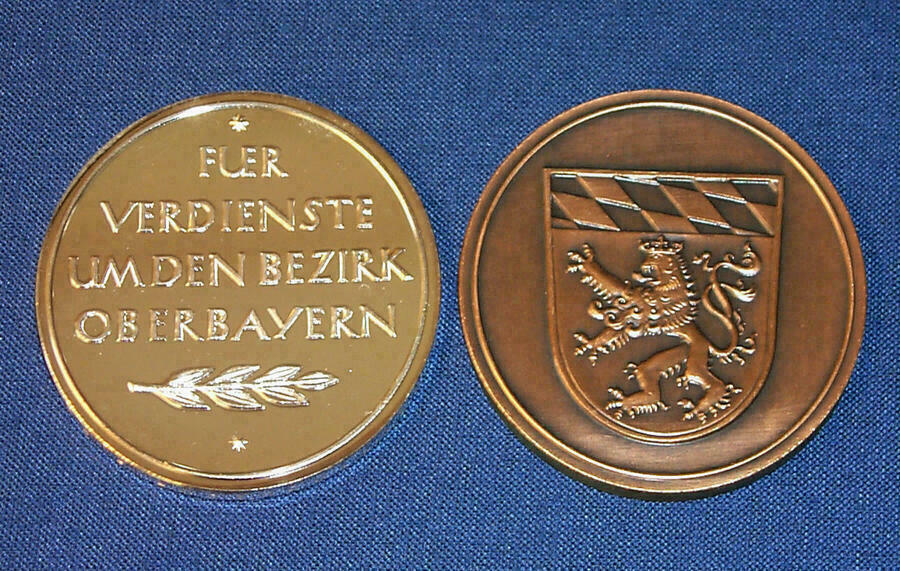 Es sind beide Seiten der Medaille zu sehen. Auf einer Seite ist der Text "Für Verdienste um den Bezirk Oberbayern" und auf der anderen Seite das Wappen des Bezirks Oberbayern eingraviert.