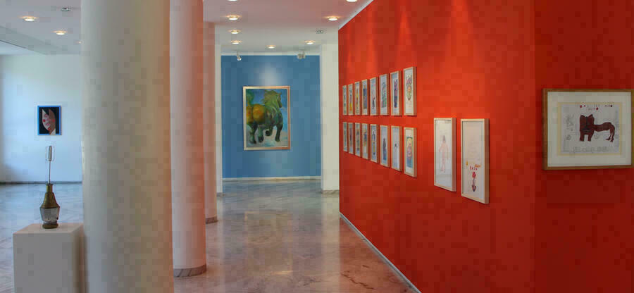 Blick in eine Ausstellung in der Galerie des Bezirks Oberbayern. An den roten und weißen Wänden gegenständliche Bilder der Künslterfamilie Hurzlmeier. Links Beton-Säulen.