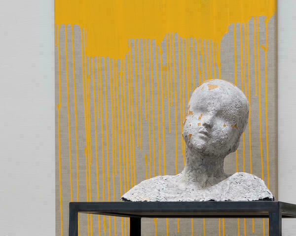 Die Skulptur eines Kopfes inklusive Schultern steht auf einem Tisch. Von einer grauen Wand im Hintergrund fließt gelbe Farbe in langen Schlieren.