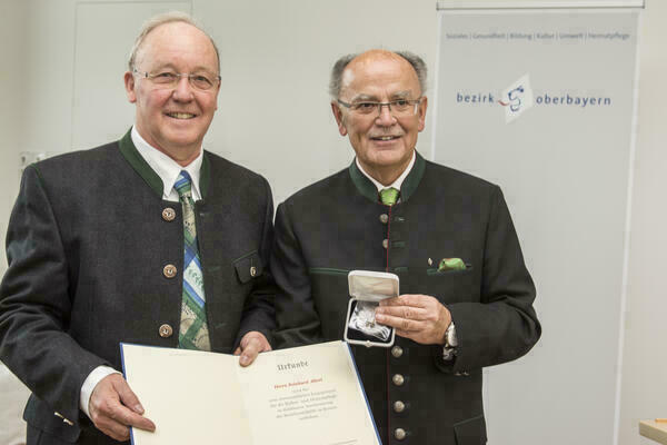 Preisträger und Bezirkstagspräsident halten lächelnd Urkunde und Medaille in die Kamera 
