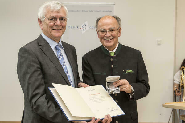 Preisträger und Bezirkstagspräsident halten lächelnd Urkunde und Medaille in die Kamera. 
