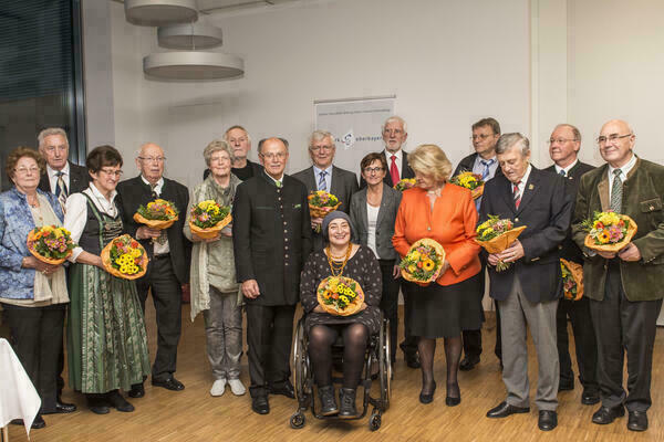 Gruppenfoto mit 16 Personen, eine Person sitzt im Rollstuhl. In der MItte steht Bezirkstagspräsident Josef Mederer. Viele halten einen Blumenstrauß in der Hand.