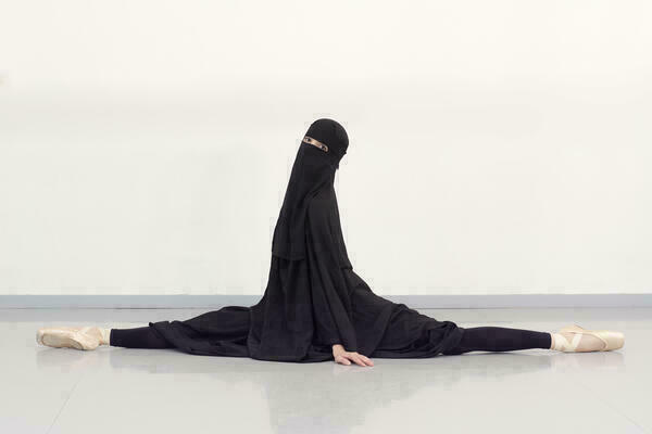 Eine Frau in Burka macht auf dem Boden einen Spagat und blickt währenddessen in die Kamera.