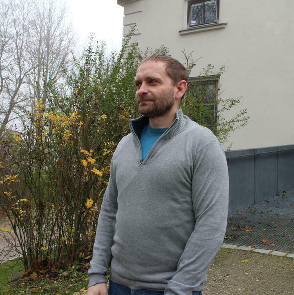Ein Mann mit grauem Pullover steht vor einem Haus. Im Hintergrund sind Büsche zu sehen.
