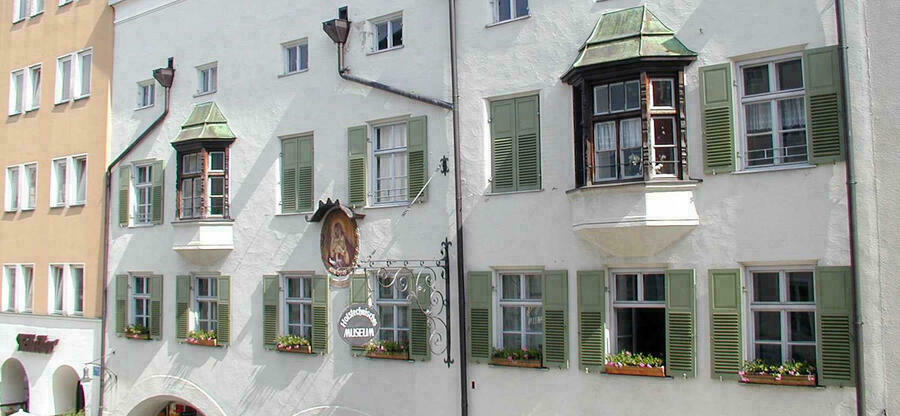 Ein weißverputztes historisches Bürgerhaus im Inn-Salzach-Stil mit Sprossen-Fenstern mit grünen Fensterläden, zwei Erkern und Regenrohren.
