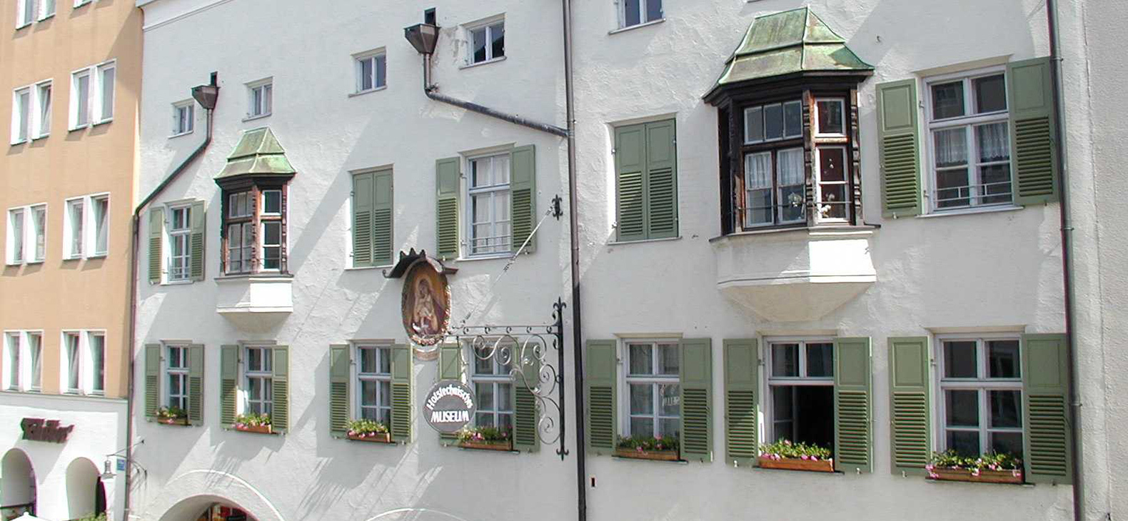 Ein weißverputztes historisches Bürgerhaus im Inn-Salzach-Stil mit Sprossen-Fenstern mit grünen Fensterläden, zwei Erkern und Regenrohren.