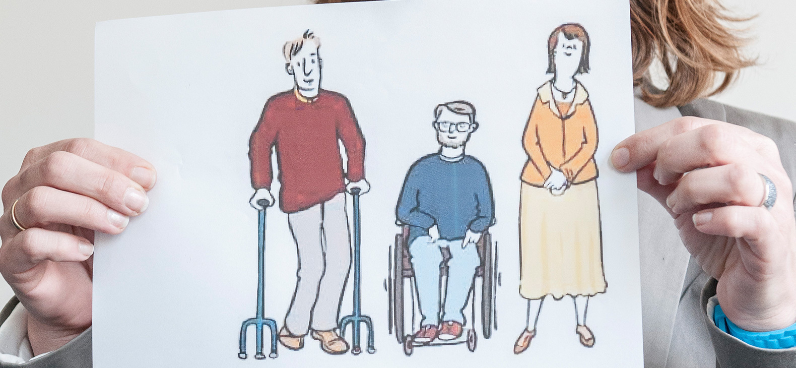 Zwei Hände halten eine Zeichnung, auf der ein Mann mit Krücken, ein Mann im Rollstuhl und eine Frau zu sehen sind.