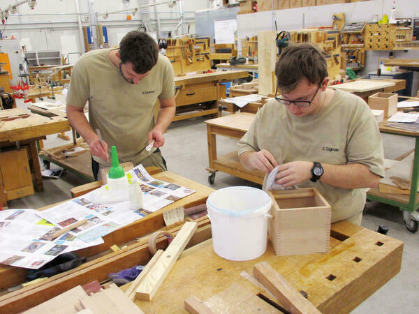 Zwei junge Männer an Werkbänken bauen Kästen aus Holz zusammen