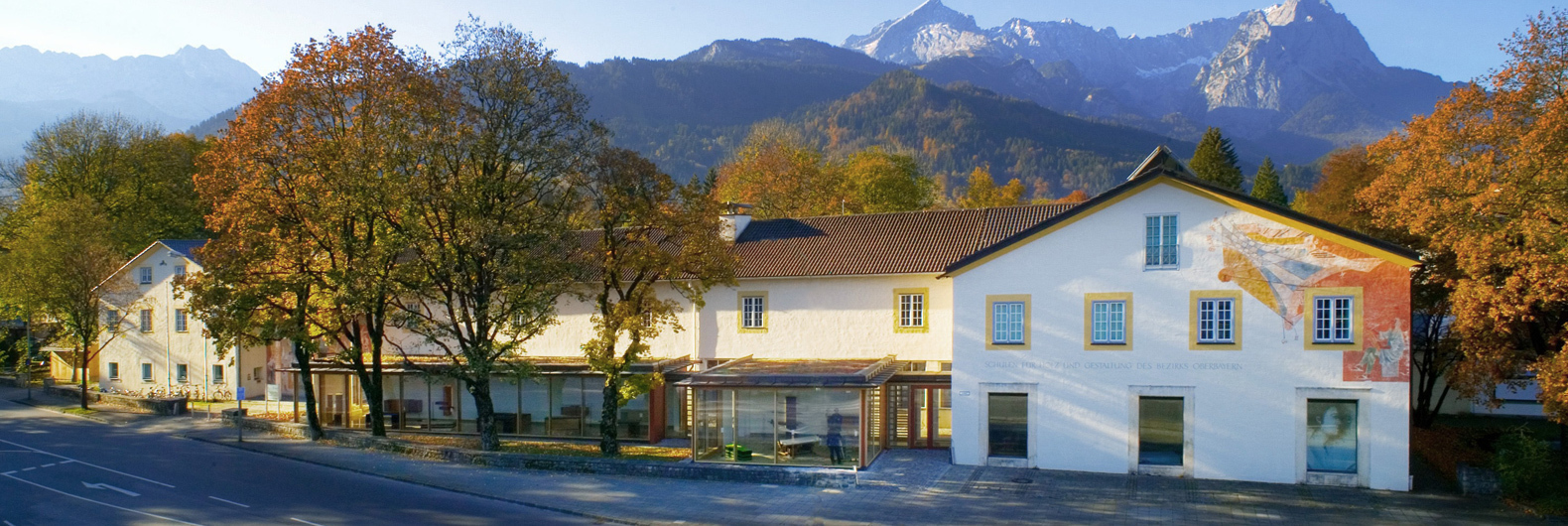 Außenansicht der Schulen für Holz und Gestaltung in Garmisch-Partenkirchen im Herbst: ein langgezogenes Gebäude mit Dreiecksgiebel und bemalter Fassade, davor Bäume und eine Straße. Im Hintergrund sieht man die Berge.