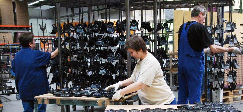 Blick in eine Werkhalle:  Eine Frau mit weißen Handschuhen handiert mit Werkstücken aus Kunststoff an einem Tish. Dahinter nehmen eine Mann und eine Frau in blauen Arbeitskleidung die gleichen Werkstücke in Hängeregale.