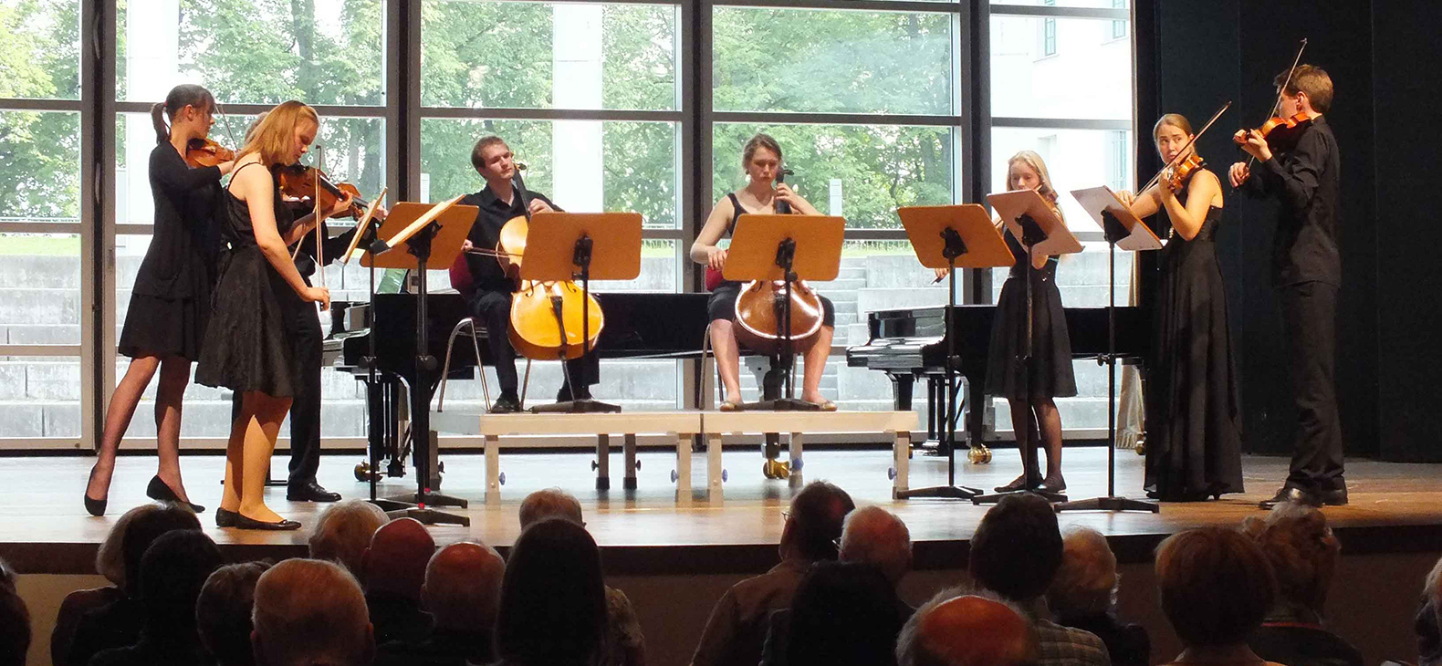 Ein Streich-Orchester mit jungen schwarz-gekleideten Musikerinnen und Musikern spielt auf einer Bühne vor einer Glasfront. Im Vordergrund erkennt man die Köpfe der Zuhörer