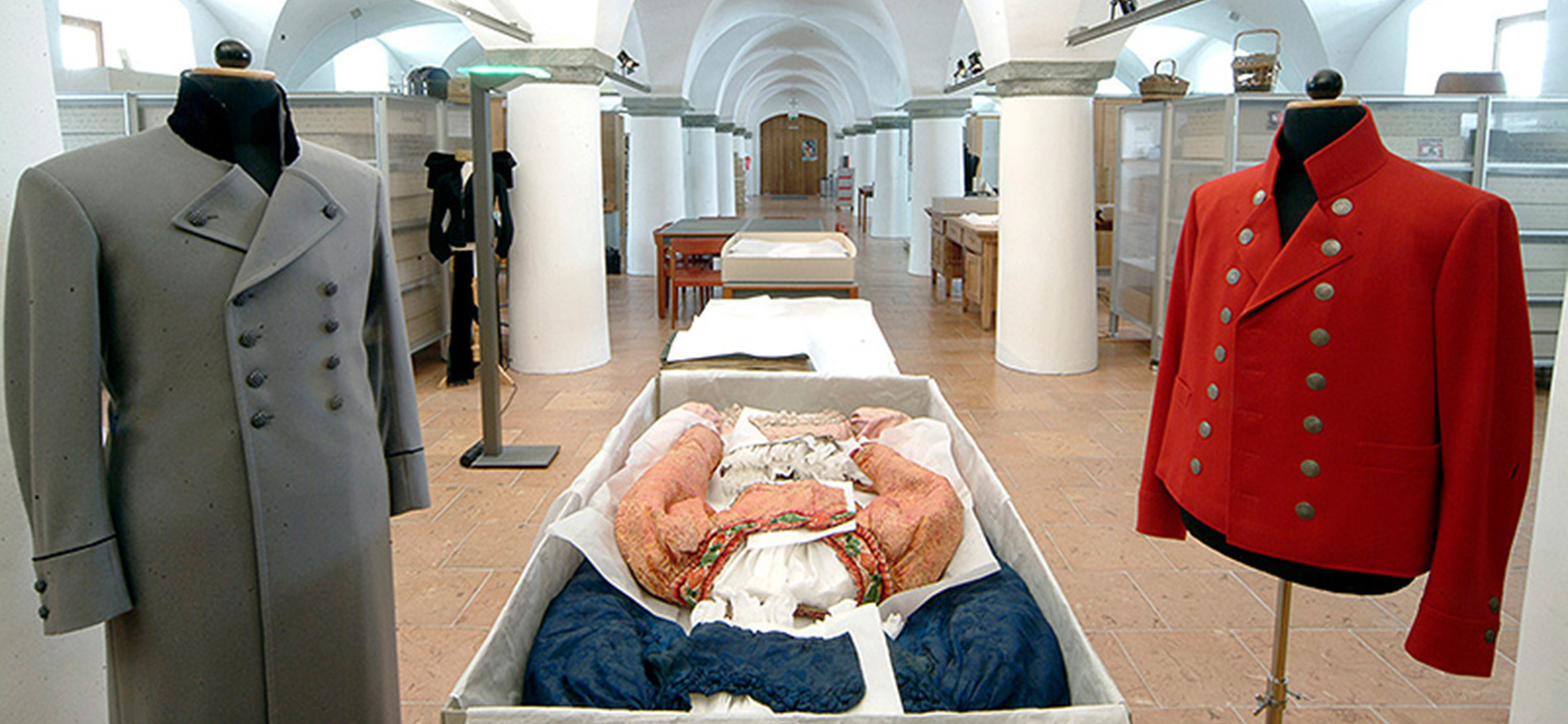 Blick in das Zentrum für Trachtengewand des Bezirks Oberbayern: Ein Raum mit kräftigen Säulen, die ein böhmisches Gewölbe tragen. Im Vordergrund sind historische Trachten ausgestellt.