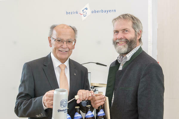 Ein Foto vom Bezirkstagspräsidenten Josef Mederer, der einen Bierkrug und vier Flaschen Bier hält, zusammen mit Bezirksrat Andreas Huber.