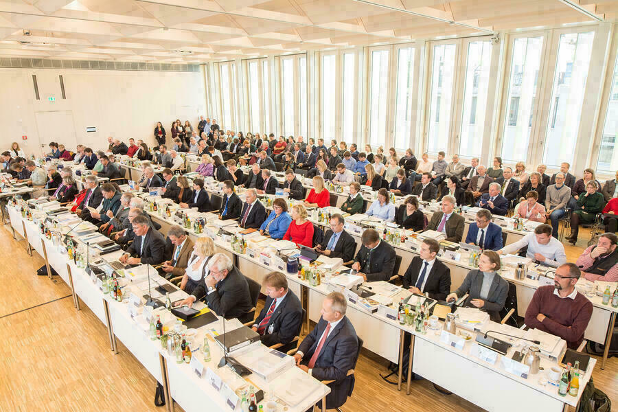 Vier Sitzreihen mit Bezirksräten im Plenarsaal des Bezirks Oberbayern. Dahinter eine Reihe mit Besucherinnen und Besuchern.