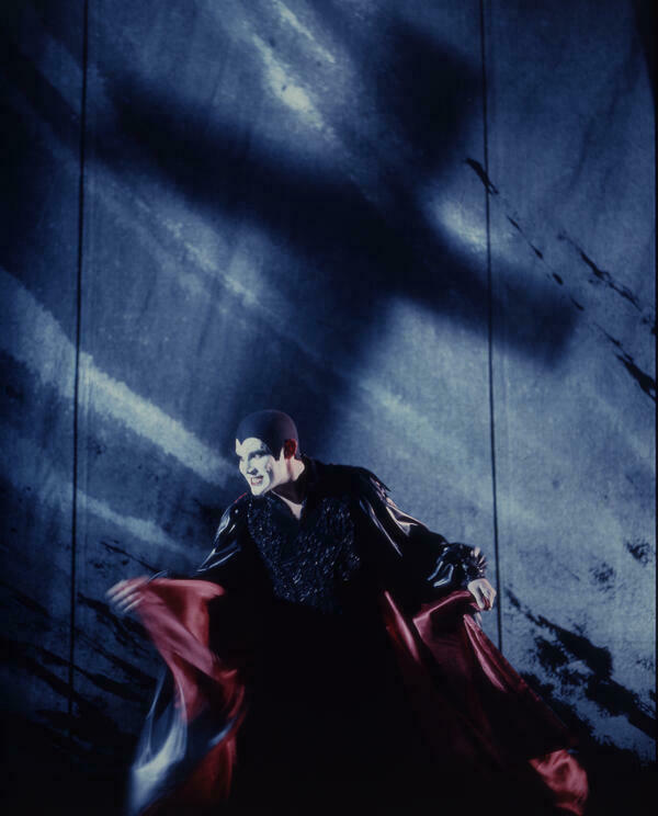 Ein Mann in einem dunklen Mephisto-Kostüm steht bösartig grinsend in einer düsteren Szenerie vor dem Schattenwurf eines überdimensionierten Kreuzes.