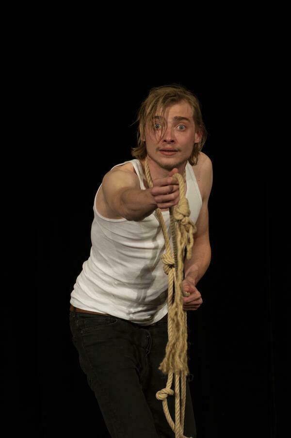 Ein junger Mann spielt im Unterhemd mit Strick um den Hals und auf den Betracher deutend auf einer Bühne vor schwarzem Hintergrund