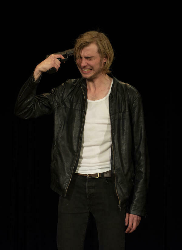 Ein junger Mann spielt in schwarzer Lederjacke mit einer Pistole in seiner Hand auf einer Bühne vor schwarzem Hintergrund