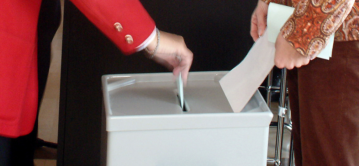 Ein Mensch wirft einen Stimmzettel in eine Wahlurne, ein anderer hält die Abdeckung in seinen Händen; es sind nur die Unterkörper zu sehen.