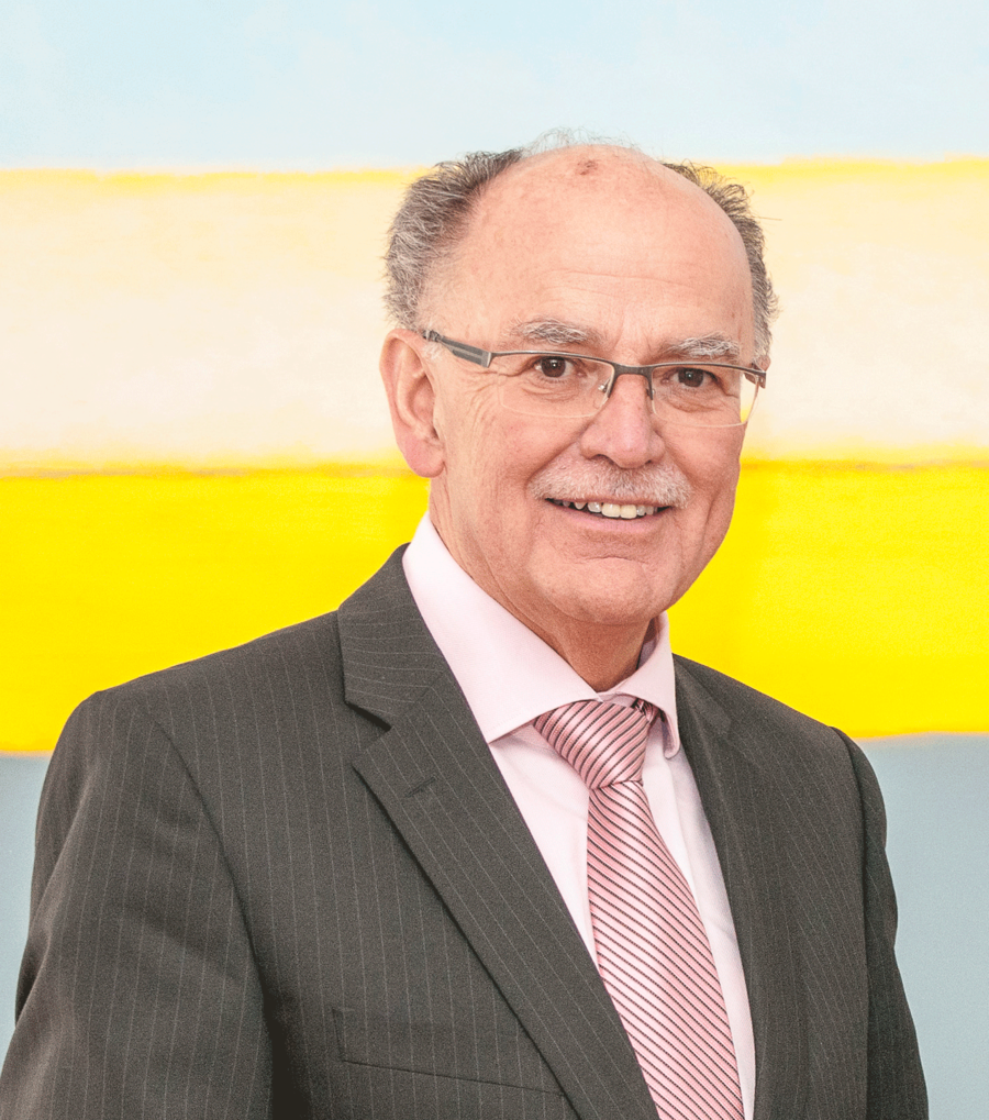 Der Bezirkstagspräsident Mederer steht vor einer Wand mit einem Gemäldein den Farben Gelb und Grau. Er trägt einen brauenen Anzug und lächelt in die Kamera.