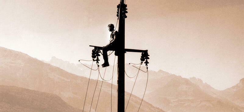 Ein Mann sitzt auf einem Strommast. Im hintergrund sieht man Gebirgszüge.