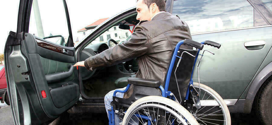 Mann sitzt im Rollstuhl vor der offenen Fahrertür eines Autos.