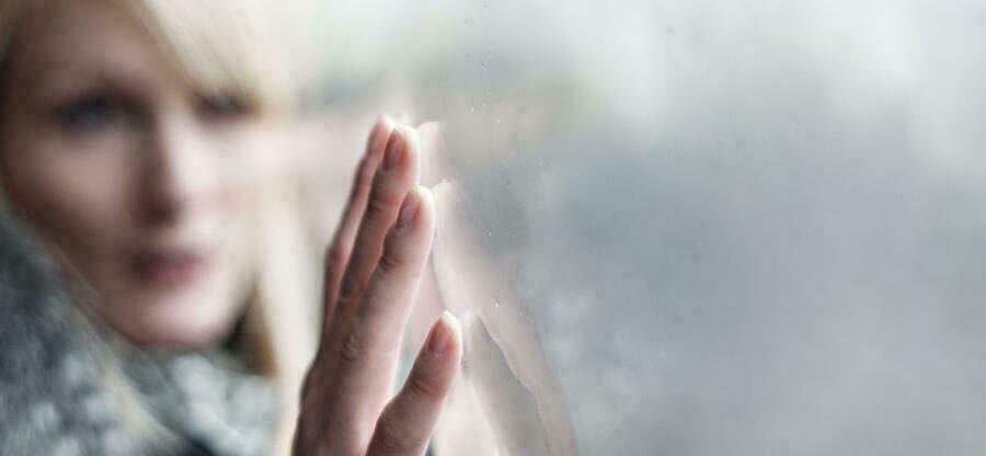 Eine Frau fährt mit ihren Fingerspitzen über eine graue Fensterscheibe. Das Gesicht der Frau ist unscharf.