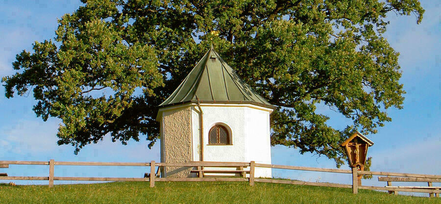Eine Kapelle und ein Wegkreuz auf einem Hügel vor einem Baum und blauem Himmel.