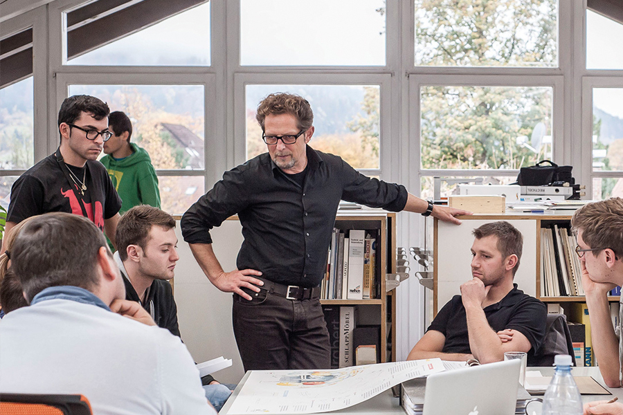 Blick in eine Klasse der Fachakademie für Raum- und Objektdesign in Garmisch-Partenkirchen:  Neun Männer sitzen und stehen vor einem großen Glasfenster an einem Tisch mit Plänen und beraten sich.