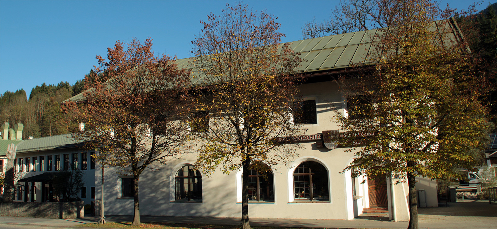 Außenansicht der Berufsfachschule für Holzschnitzerei und Schreinerei in Berchtesgaden: ein traditionelles Gebäude mit Spitzgiebel und Rundbogenfenstern, davor mehrere Bäume.