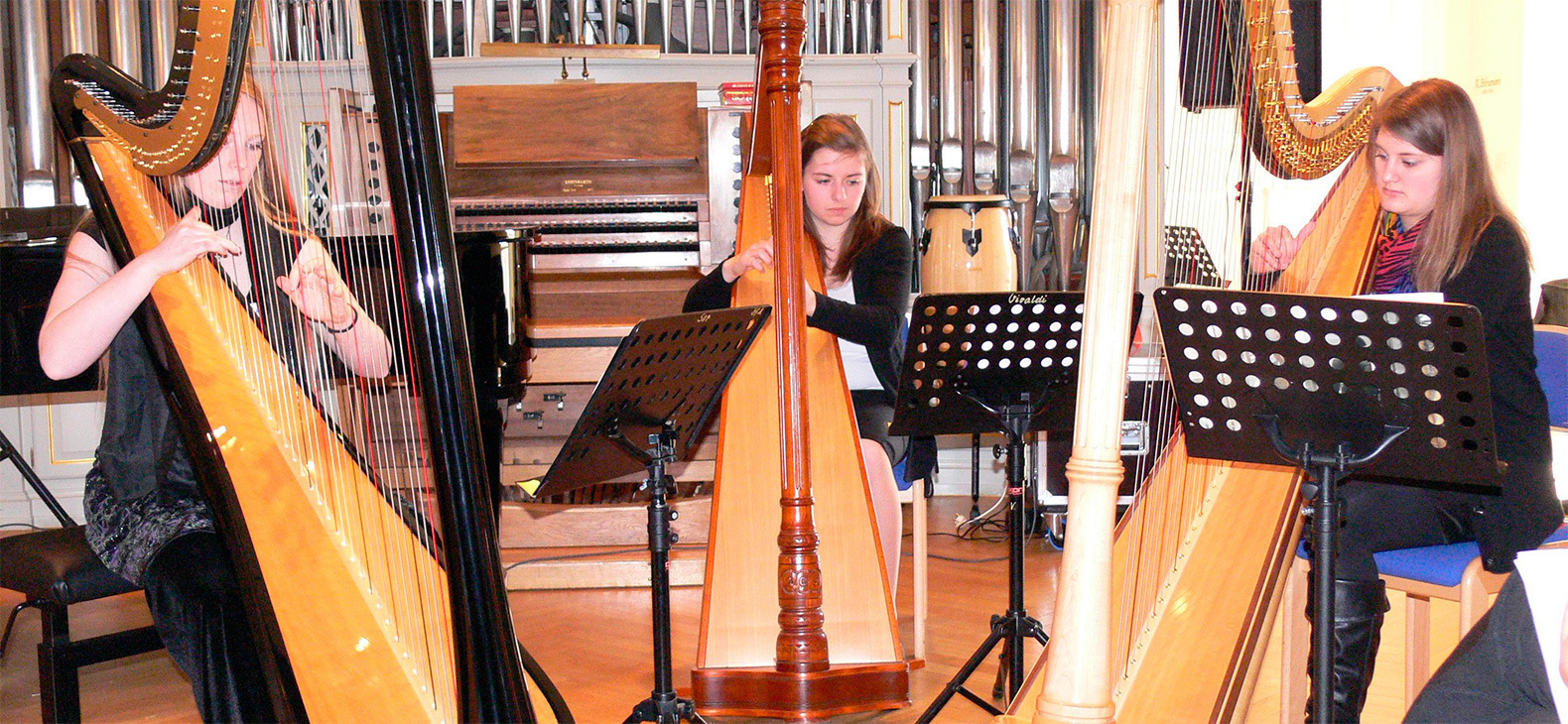 Drei Frauen spielen Harfe. Im Hintergrund ist eine Orgel zu erkennen.