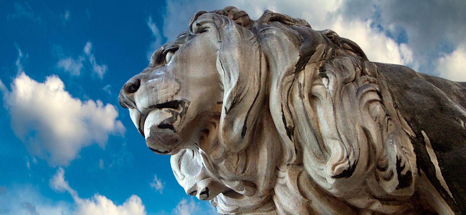 Eine Löwenskulptur schaut in die Ferne, darüber ein blauer HImmel mit Wolken