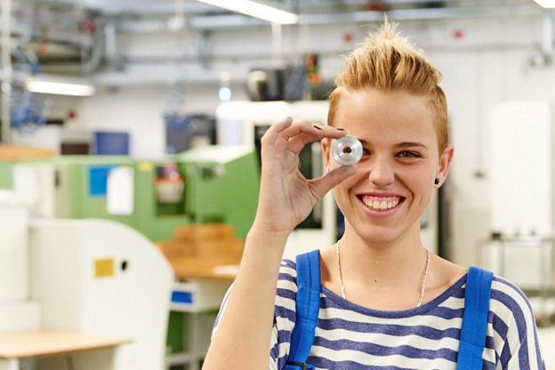Eine lachende junge Frau mit kurzen blonden Haaren und einer blauen Arbeitshose hält einen Metallring vor das rechte Auge. Im Hintergrund sieht man eine Werkhalle.