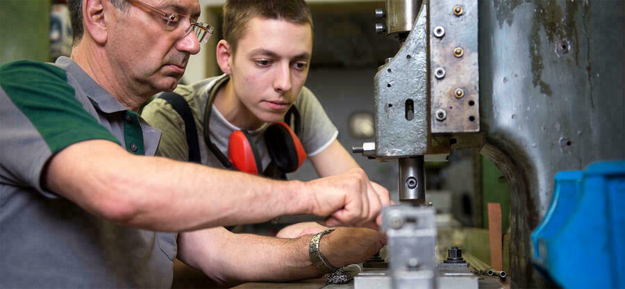 Ein junger Mann sieht einem lteren Mann aufmerksam dabei zu, wie er an einer Maschine zur Metallverarbeitungan einem Werkstck arbeitet.