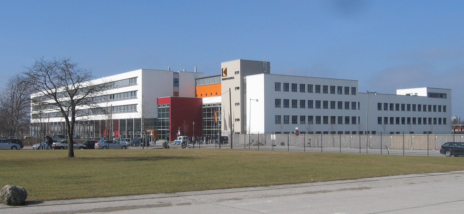 Außenansicht der Adolf-Kolping-Berufsschule in München:
Blick von einer Straße über einen Rasen auf einen Gebäude-Komplex aus Längs- und Querriegeln.