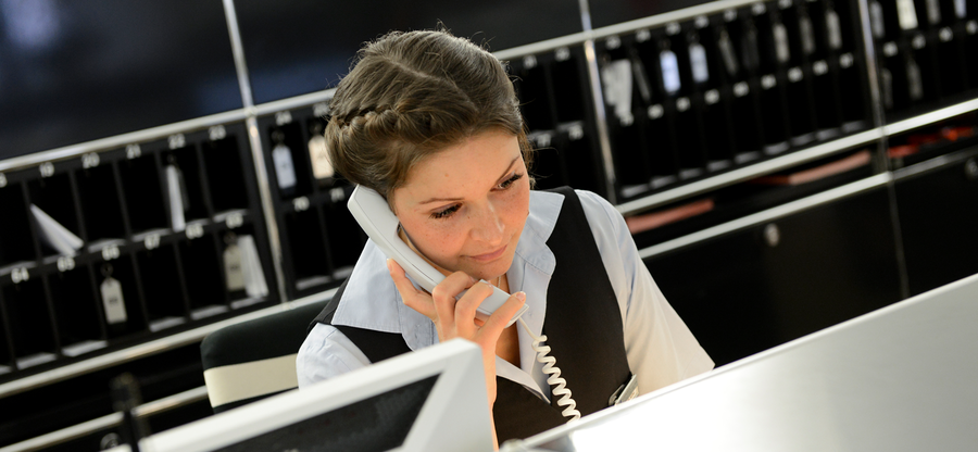 Eine junge Frau mit heller Bluse und dunkelblauer Weste sitzt an einer Hotel-Rezeption und telefoniert. Sie trägt ihr Haar nach oben geflochten.