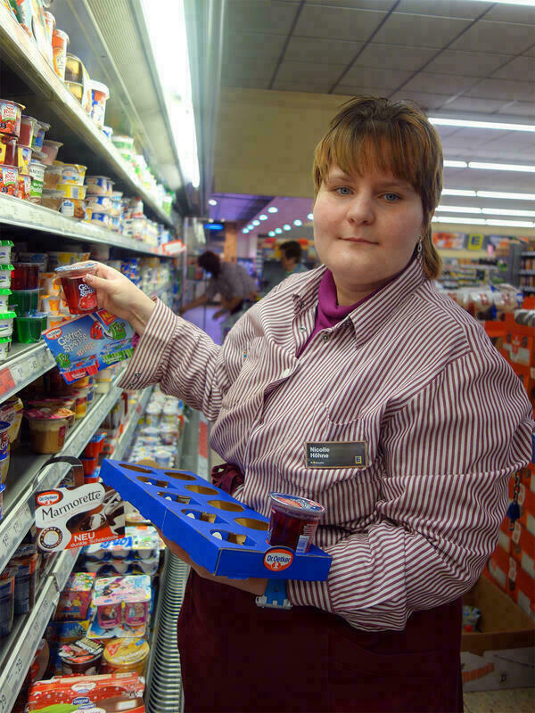 Eine Frau räumt das Kühlregal im Supermarkt ein und schaut dabei in die Kamera.