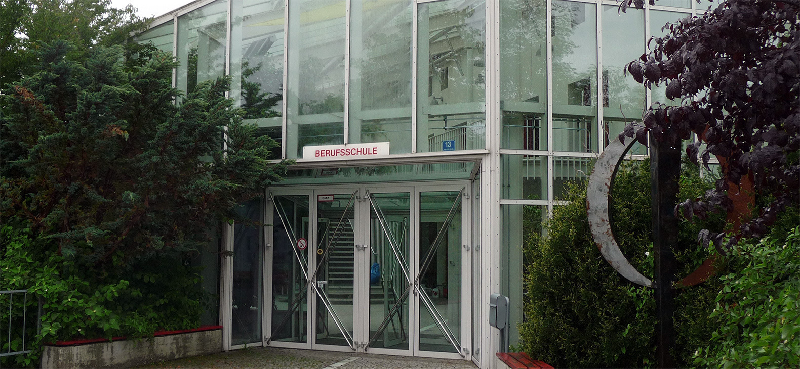 Eingang zur Albrecht-Schnitter-Schule: Eine zweigesschossige Glasfront wird rechts und links von hohen Büschen eingerahmt.