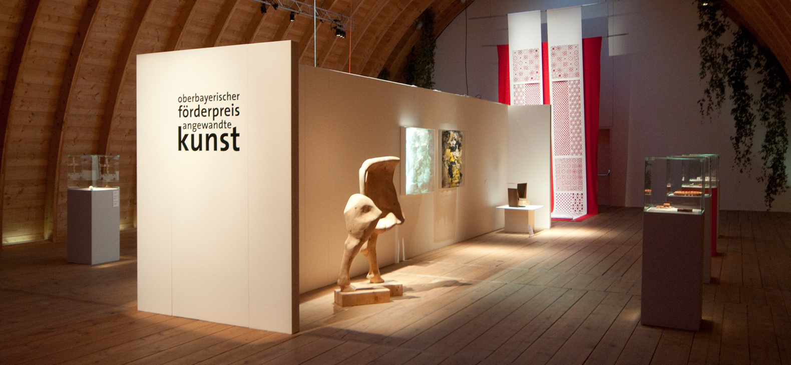 Blick in die Ausstellung, es sind mehrere Kunstwerke zu sehen. In der MItte eine Trennwand mit dem Schriftzug "oberbayerischer  förderpreis angewandte kunst" am Kopfende.